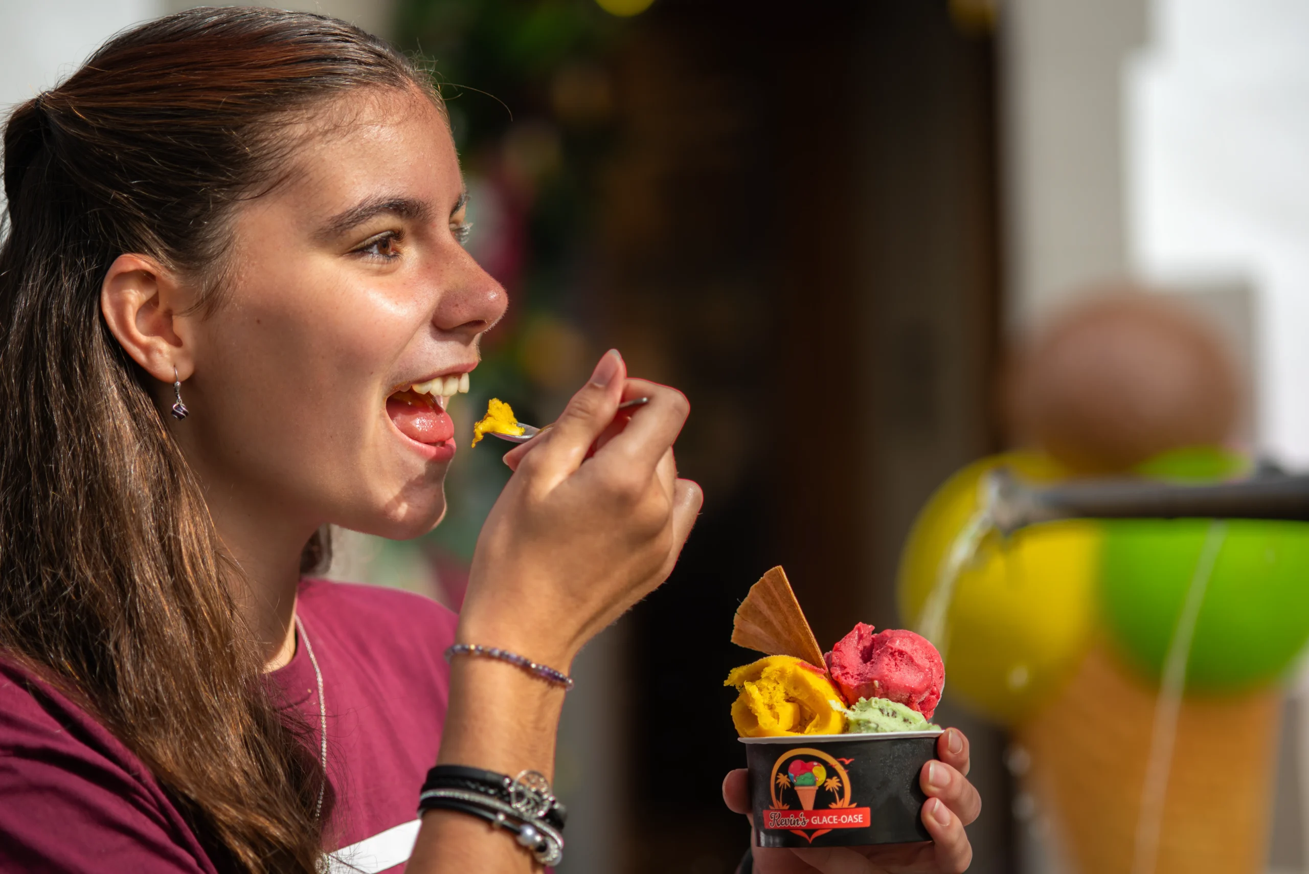 Junge Frau mit zusammengebundenen braunen Haaren laechelt und isst Eis aus einem Becher mit einem Loeffel waehrend sie draussen steht.