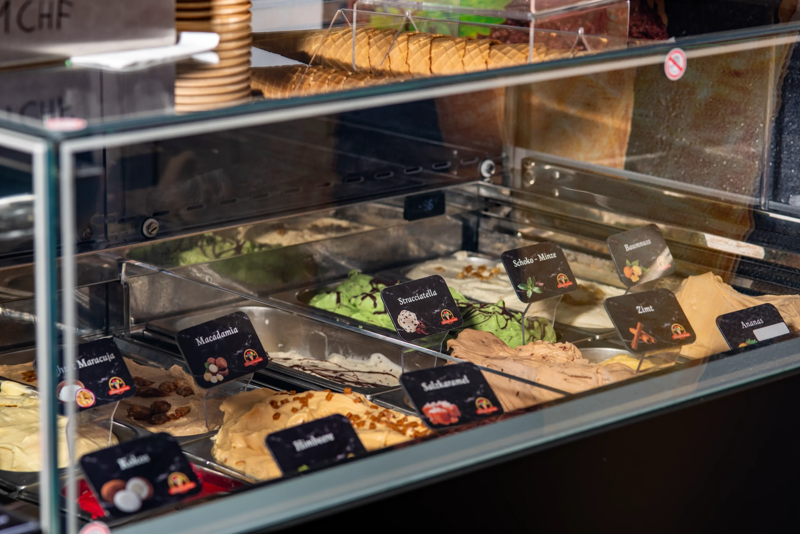 Eine Eistheke mit vielen Sorten in einer Vitrine, beschriftet und frisch angerichtet, dazu Waffeln im Hintergrund.