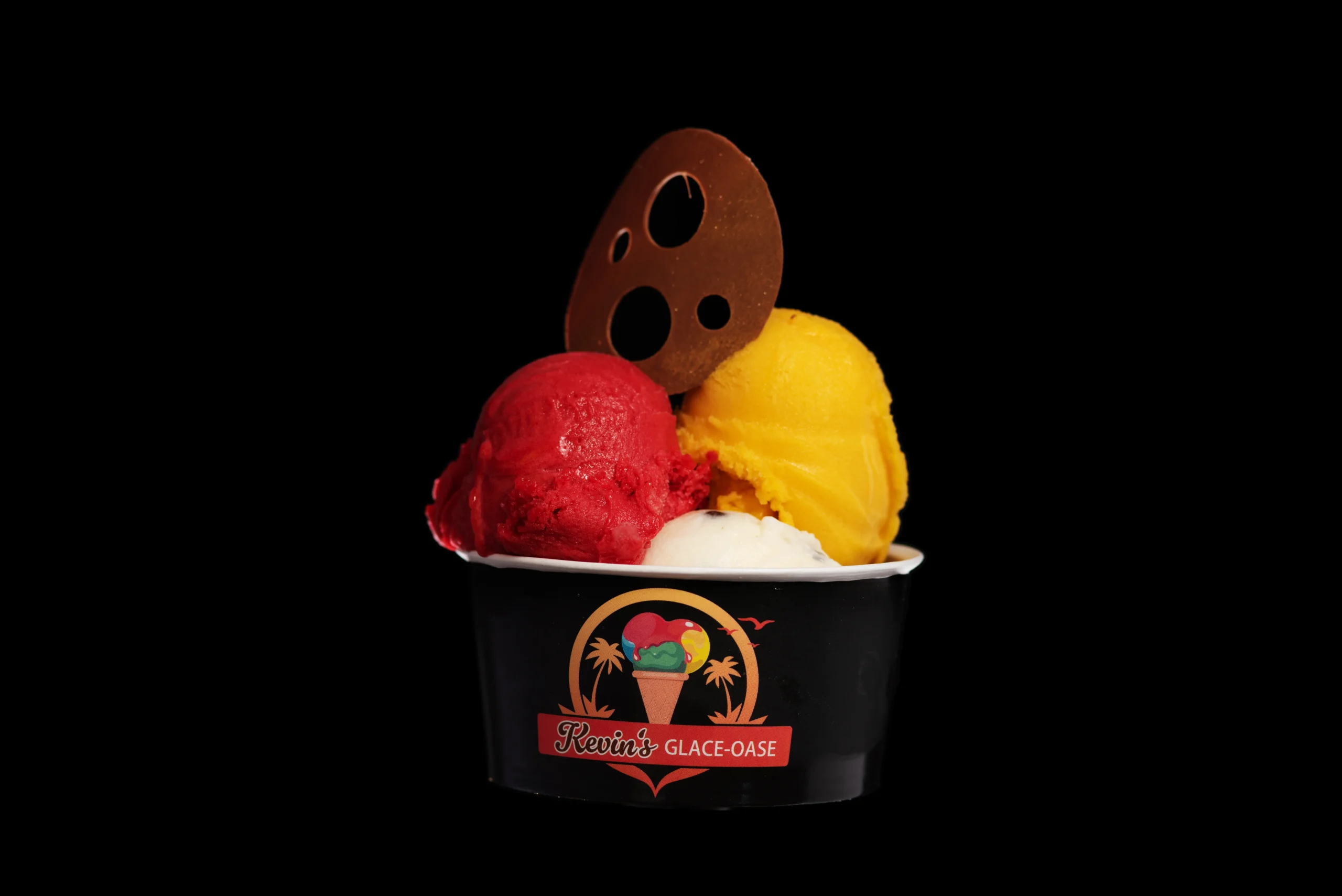 Becher mit drei Kugeln Eis (rot, gelb, weiß) und Schokoladendeko vor schwarzem Hintergrund.