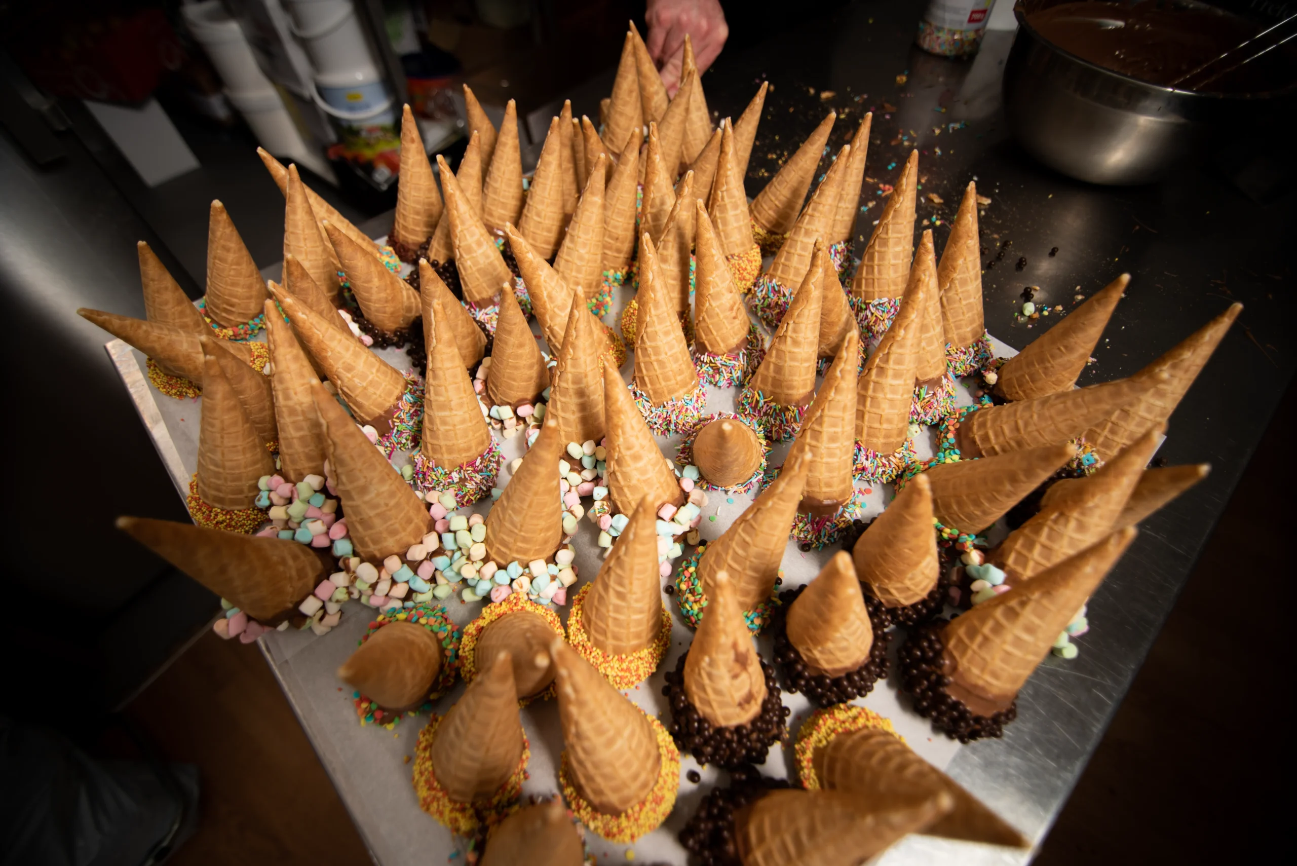 Viele dekorierte Eiswaffeln stehen kopfüber auf einem Tisch, umrandet von Schokolade, Streuseln und Mini-Marshmallows.