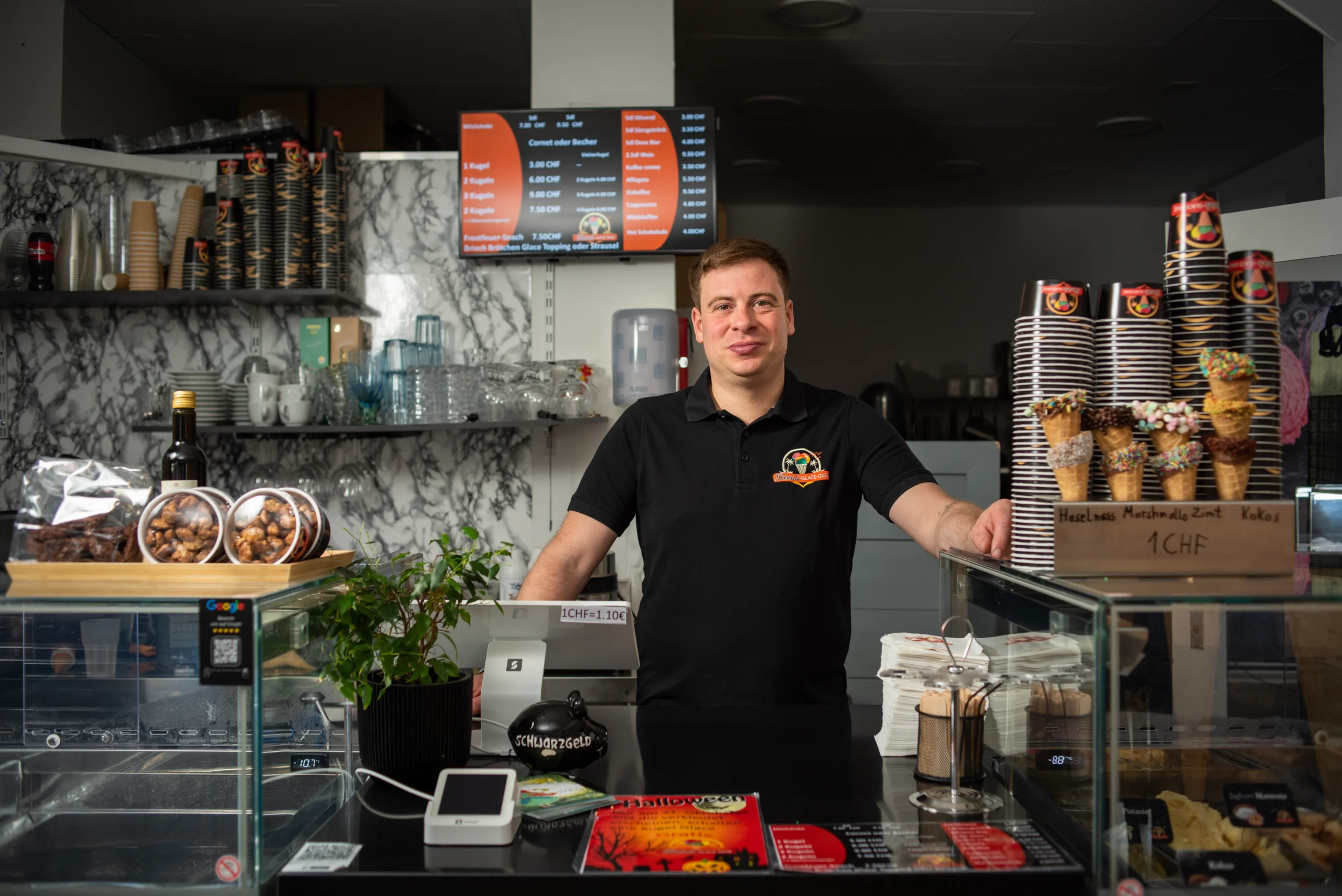 Ein Mann steht hinter der Theke einer Eisdiele mit Eiswaffeln Vitrinen und Preistafel im Hintergrund.